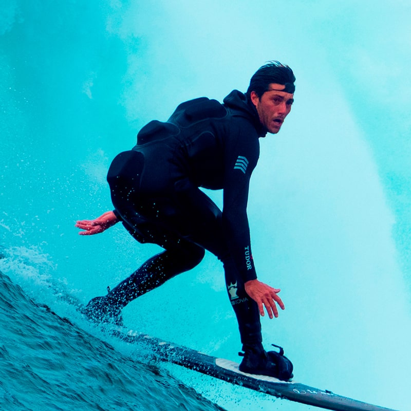 Ein Mann in einem schwarzen Neoprenanzug surft auf einer Welle, lehnt sich nach vorne und balanciert auf seinem Brett, während hinter ihm das blaue Wasser plätschert - eine aufregende Szene, die an die abenteuerlustigen Entdecker der Tudorzeit erinnert.