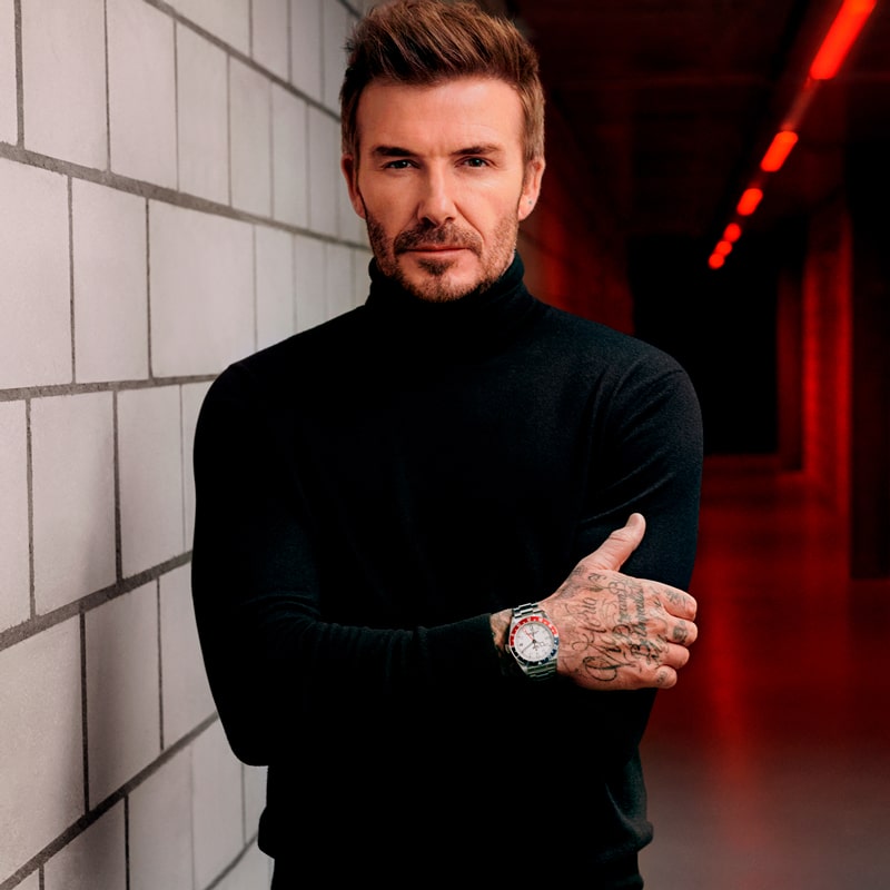 Ein Mann mit gestyltem Haar und Gesichtsbehaarung, der einen schwarzen Rollkragenpullover und eine Tudor-Uhr trägt, steht an einer gekachelten Wand in einem schwach beleuchteten Korridor mit roten Lichtern. Er hat Tätowierungen an der Hand und blickt in die Kamera.