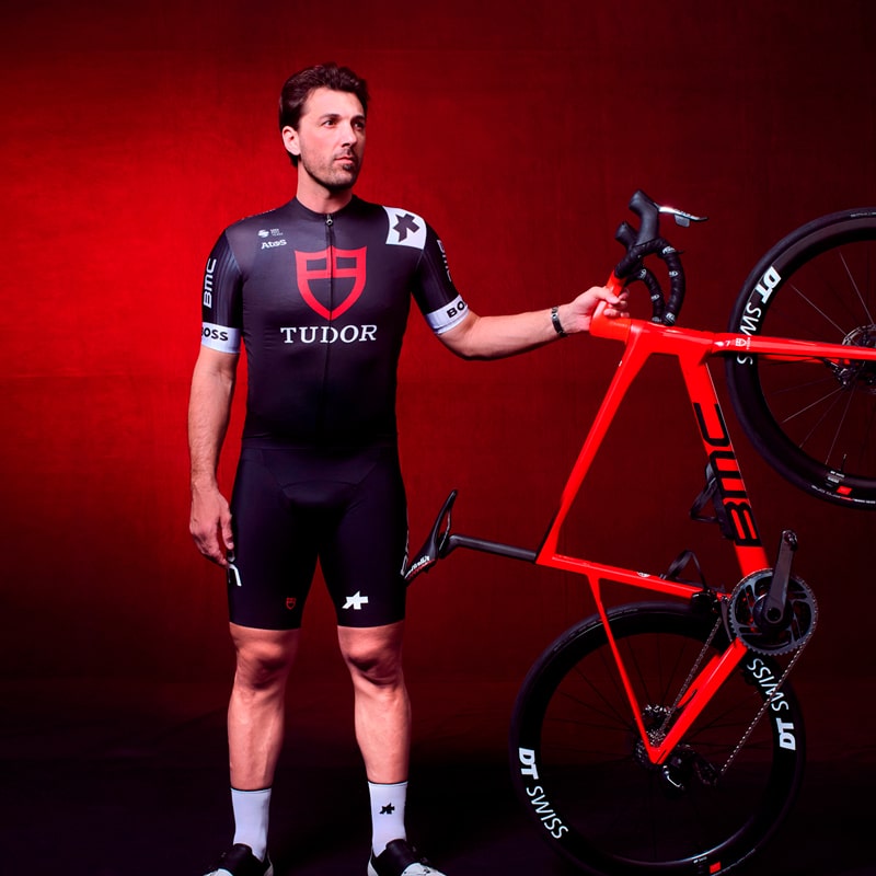 Ein Tudor-Radfahrer in schwarzer Teamkleidung steht vor einem roten Hintergrund und hält ein rotes BMC-Rennrad mit einer Hand am Lenker aufrecht.