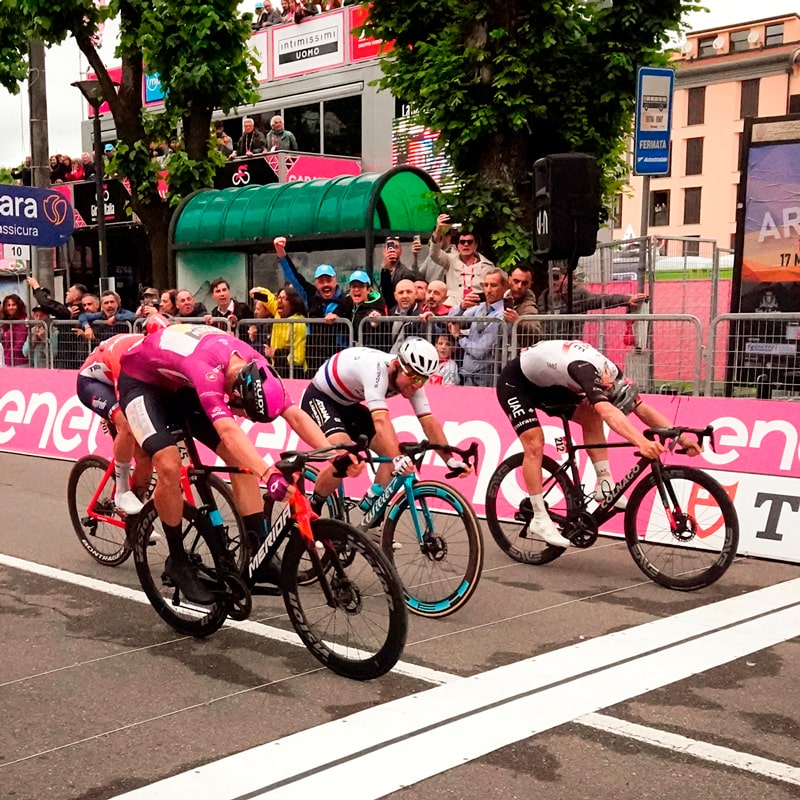 Vier Radfahrer rasen auf ihren Tudor-Rädern nach vorne gelehnt auf die Ziellinie zu. Zuschauer jubeln ihnen hinter einer Absperrung zu, und Veranstaltungsbanner und Bäume säumen die Straße. Die Atmosphäre ist intensiv und wettbewerbsorientiert.