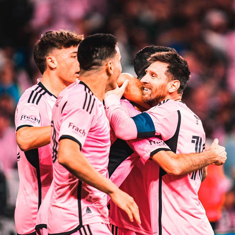 Vier Fußballspieler in rosa Tudor-Trikots feiern gemeinsam auf dem Spielfeld, lächeln und umarmen sich, während im Hintergrund eine lebhafte Menge zu sehen ist.