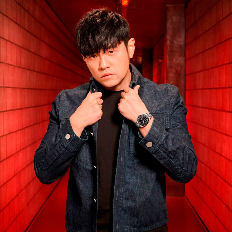 Ein Mann mit schwarzem Haar, der eine dunkle Jeansjacke im Tudor-Stil und ein schwarzes Hemd trägt, steht in einem rot beleuchteten Flur, rückt den Kragen seiner Jacke zurecht und schaut ernst in die Kamera.