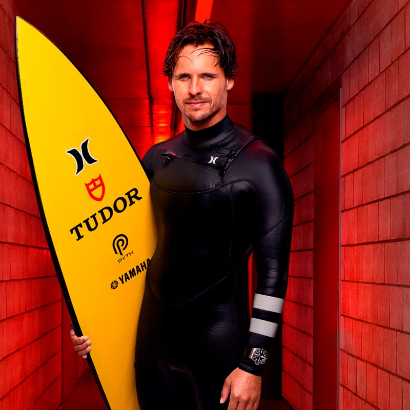 Ein Mann in einem schwarzen Neoprenanzug steht in einem rot beleuchteten Korridor und hält ein gelbes Surfbrett mit Tudor- und anderen Logos in der Hand. Mit seinem kurzen braunen Haar, der hellen Gesichtsbehaarung und einer Tudor-Uhr am linken Handgelenk strahlt er Selbstbewusstsein und Stil aus.