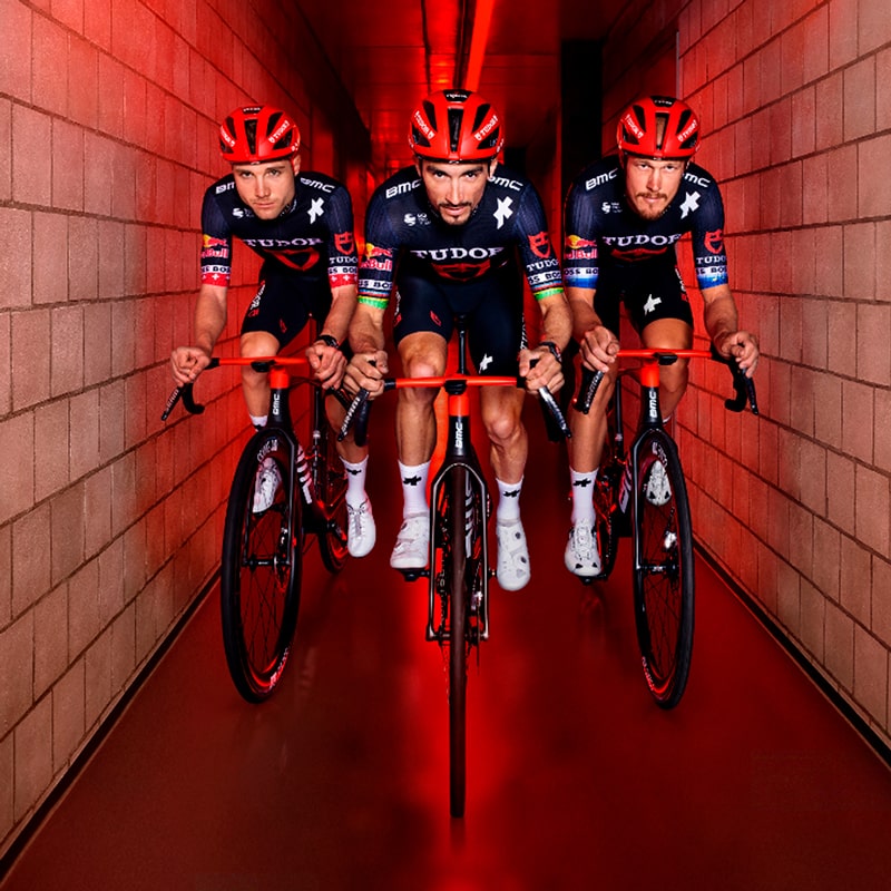 Drei männliche Radfahrer in passender schwarz-roter Tudor-Teamkleidung fahren mit ihren Fahrrädern nebeneinander einen schmalen, rot beleuchteten Gang mit Backsteinwänden hinunter und zeigen so die Einigkeit und Entschlossenheit der Tudor-Mannschaft.