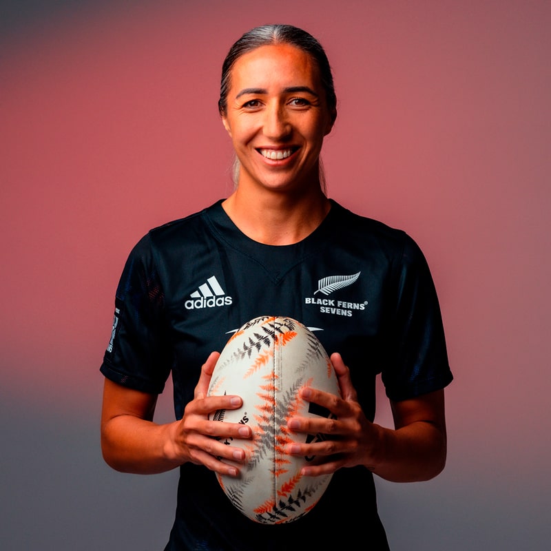 Eine lächelnde Rugbyspielerin in einem schwarzen "Black Ferns Sevens"-Trikot und einer Tudor-Uhr hält einen Rugbyball vor einem rosa Hintergrund.