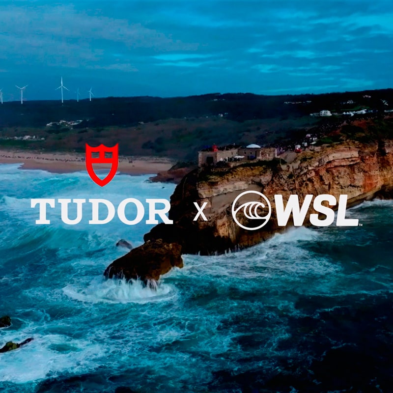 Luftaufnahme von großen Meereswellen, die gegen felsige Klippen in der Nähe eines Strandes schlagen, mit überlagerten "Tudor x WSL"-Logos. Windturbinen stehen auf den Hügeln im Hintergrund unter einem wolkenverhangenen Himmel und betonen den Abenteuergeist von Tudor.
