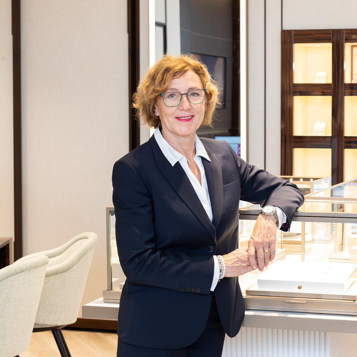 © Juwelier Wenthe Eine Frau mit kurzen lockigen Haaren und Brille, die einen marineblauen Anzug und ein weißes Hemd trägt, steht lächelnd neben einer Glasvitrine in einem modernen, gut beleuchteten Geschäft oder Ausstellungsraum.
