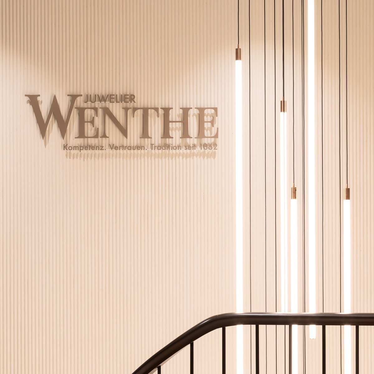 © Juwelier Wenthe Minimalistisches Interieur mit vertikalen Hängelampen neben einer Wand mit der Aufschrift "JUWELIER WENTHE" und dem Slogan "Kompetenz. Vertrauen. Tradition. Seit 1862"; ein dunkles Geländer wölbt sich nach unten.