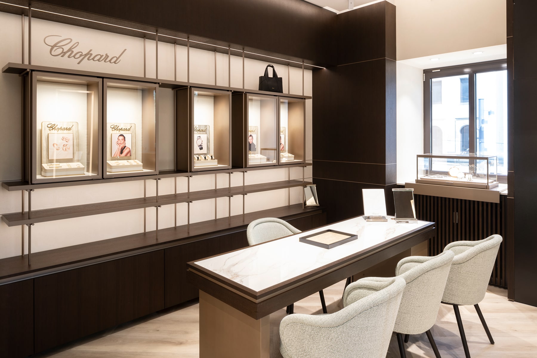 © Juwelier Wenthe Ein modernes Juweliergeschäft mit einem Beratungstisch aus Marmor, gepolsterten Stühlen, Vitrinen mit Uhren und Schmuck sowie Regalen mit dem Chopard-Logo an der Wand.