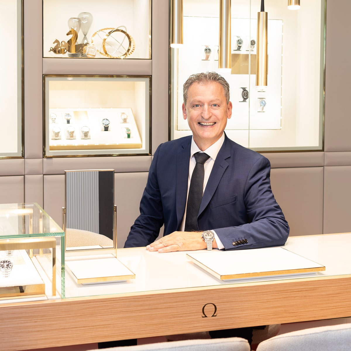 © Juwelier Wenthe Ein Mann im marineblauen Anzug und mit Krawatte sitzt lächelnd am Tresen eines Luxusuhrengeschäfts mit Uhrenvitrinen und elegantem Dekor im Hintergrund.