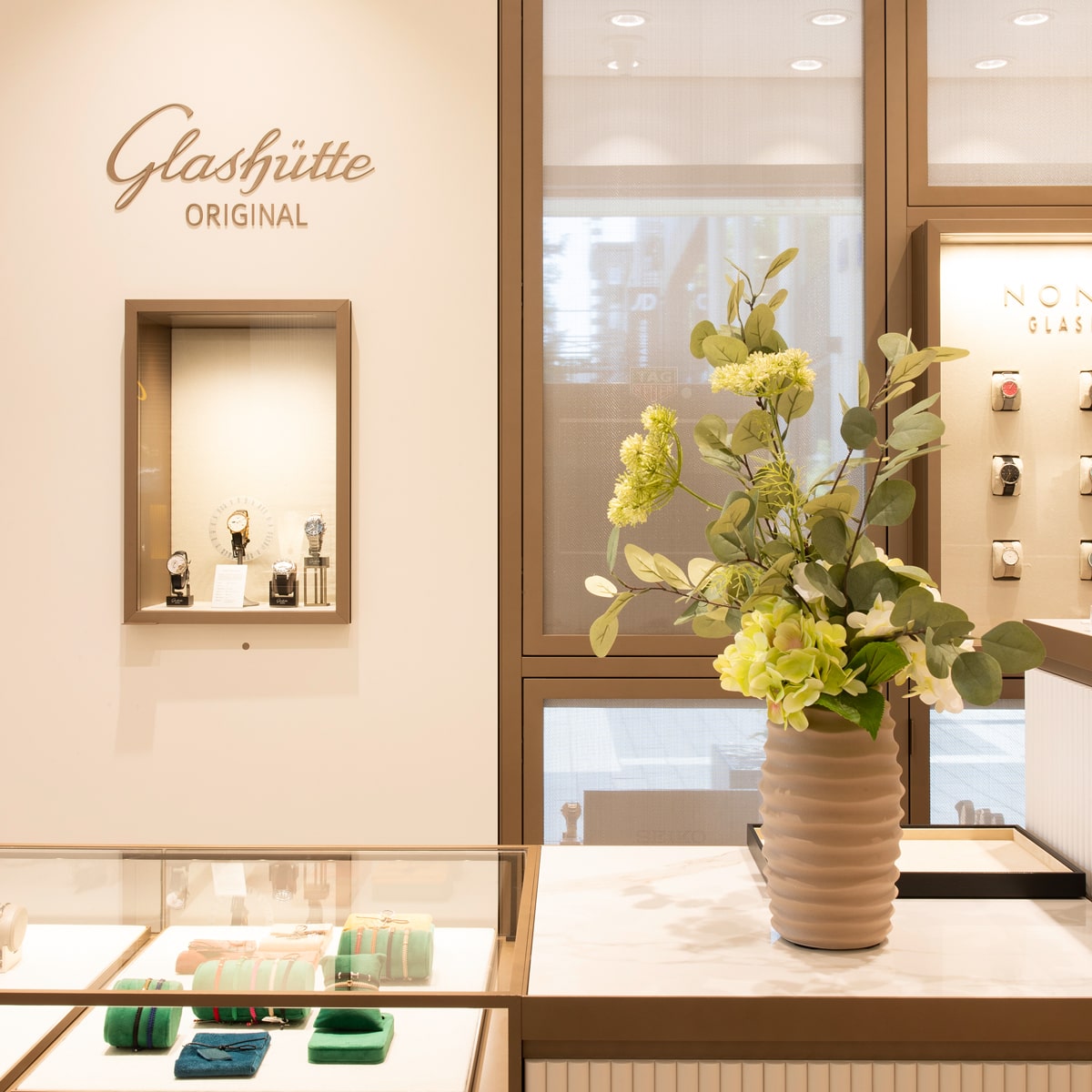 © Juwelier Wenthe In einer hellen, eleganten Uhrenboutique von Glashütte Original sind Luxusuhren in Vitrinen und an der Wand ausgestellt. Eine beigefarbene Keramikvase mit gelben Blumen steht auf einem Tresen neben großen Fenstern.