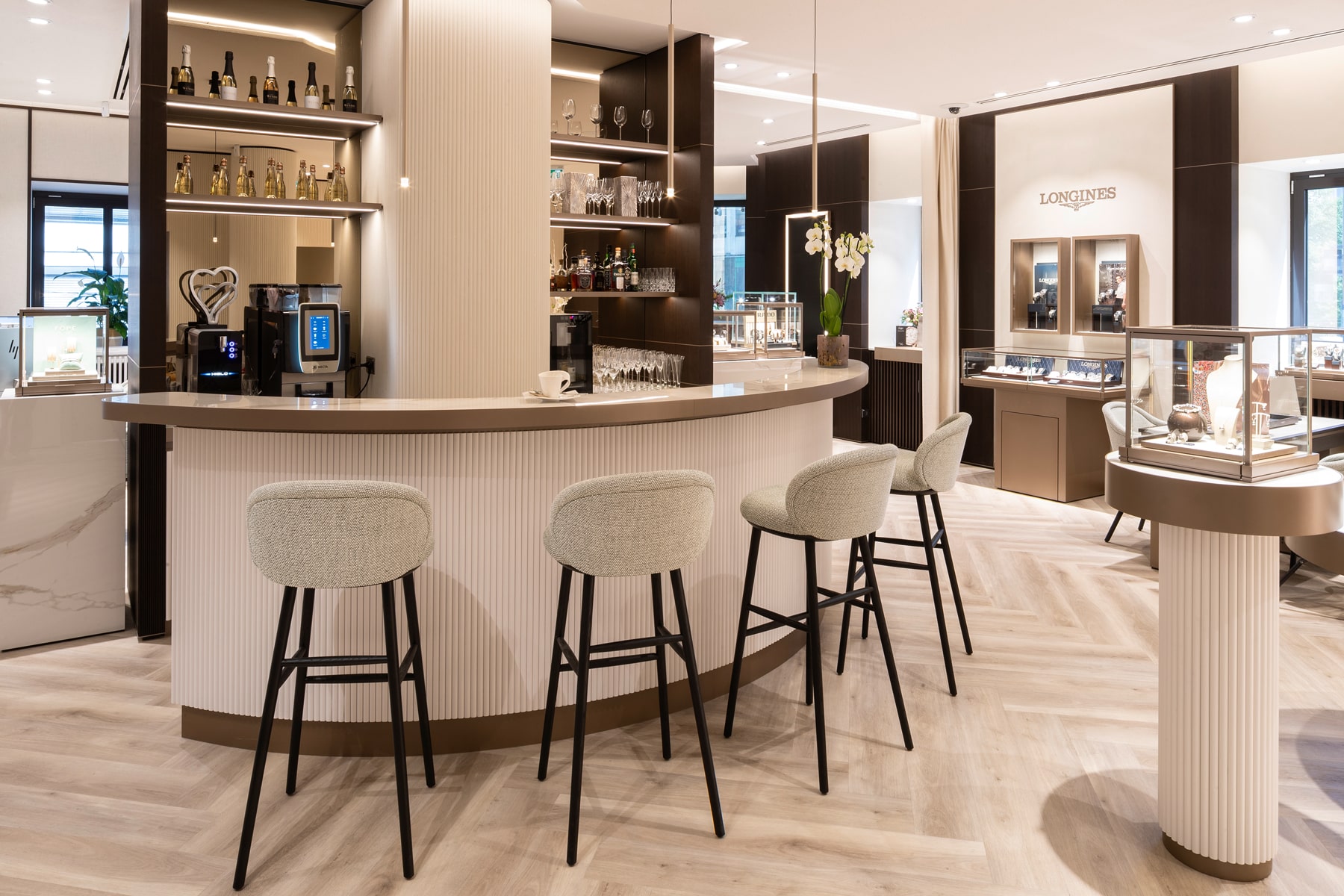 © Juwelier Wenthe Moderner, eleganter Barbereich mit einem geschwungenen Tresen, vier gepolsterten Barhockern, Glasregalen mit Flaschen und Gläsern sowie Vitrinen mit Uhren in einem hellen, gehobenen Verkaufsraum.