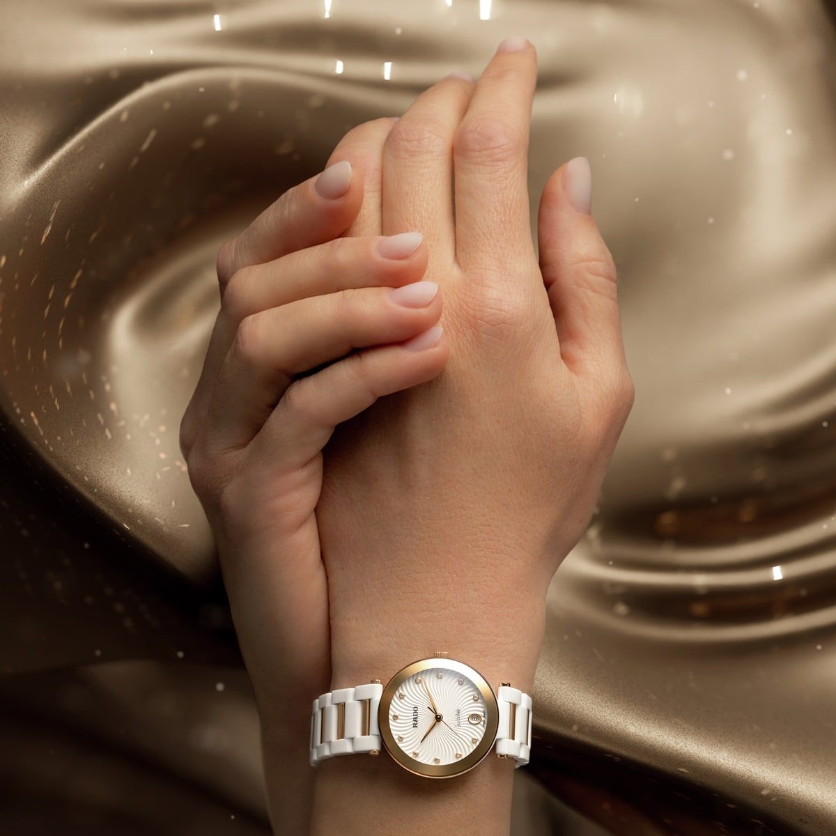 Eine Nahaufnahme von zwei sanft aneinandergelegten Händen, die eine weiß-goldene Armbanduhr von Rado LaCoupole tragen, vor einem schimmernden, goldenen, seidigen Stoffhintergrund.