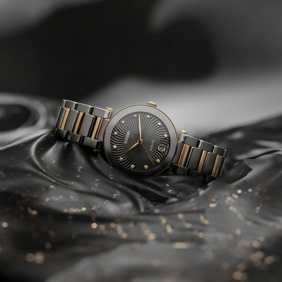 Eine luxuriöse Rado LaCoupole-Armbanduhr mit schwarzem und goldenem Metallband ruht auf schwarzem Seidenstoff mit goldenen Flecken. Das Zifferblatt zeigt ein kompliziertes, wirbelndes schwarzes Muster mit goldenen Akzenten und ein kleines Datumsfenster.