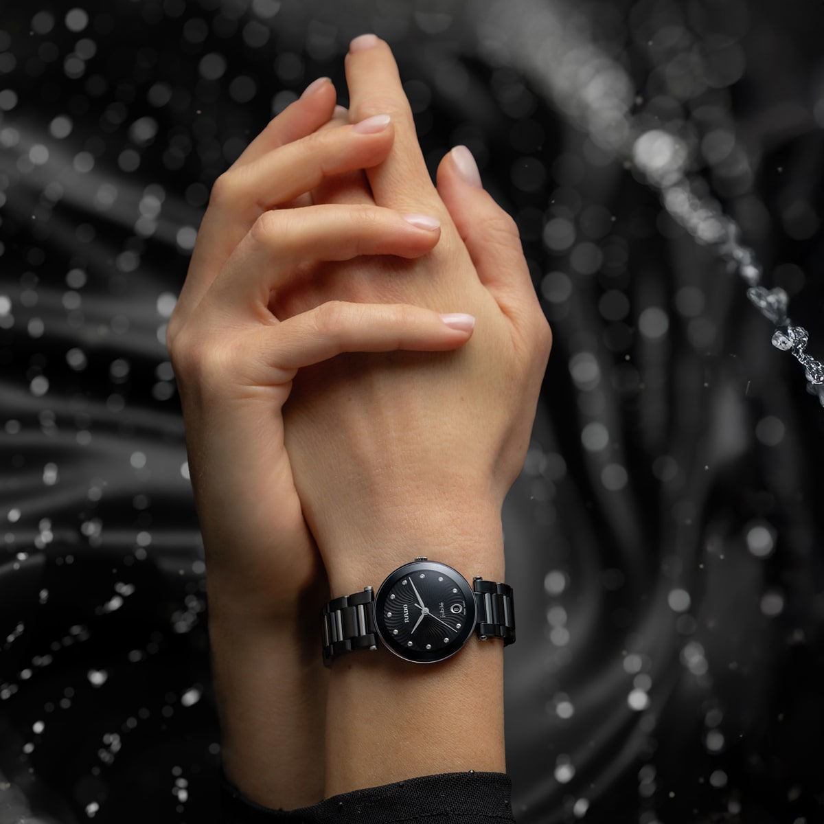 Nahaufnahme von zwei ineinander verschränkten Händen, eine davon mit einer eleganten Rado LaCoupole Armbanduhr, während Wassertropfen vor einem dunklen, unscharfen Hintergrund glitzern und einen dramatischen Effekt erzeugen.