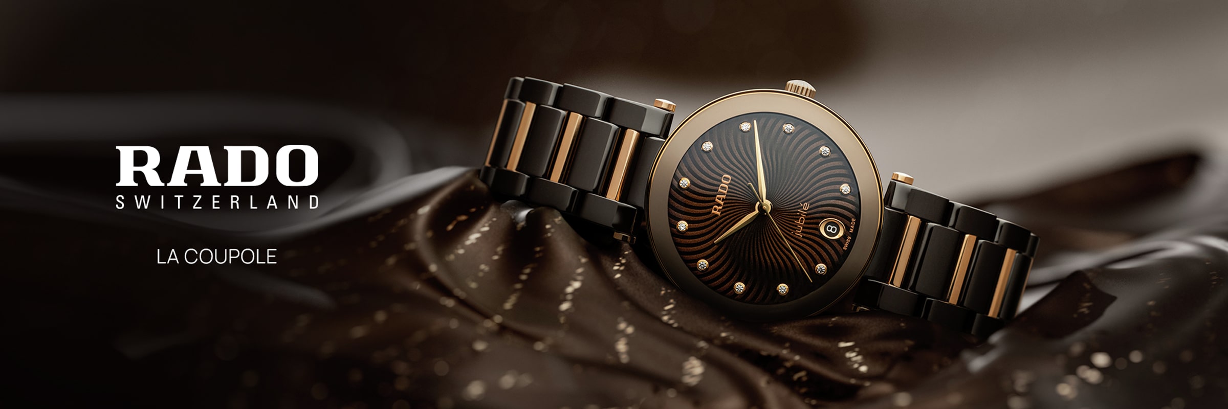 Eine Rado LaCoupole-Armbanduhr mit schwarz-goldenem Design ruht auf einem glänzenden, gemusterten Stoff. Das Zifferblatt ist mit goldenen Akzenten und diamantbesetzten Indexen versehen, während auf der linken Seite "RADO SWITZERLAND" steht.