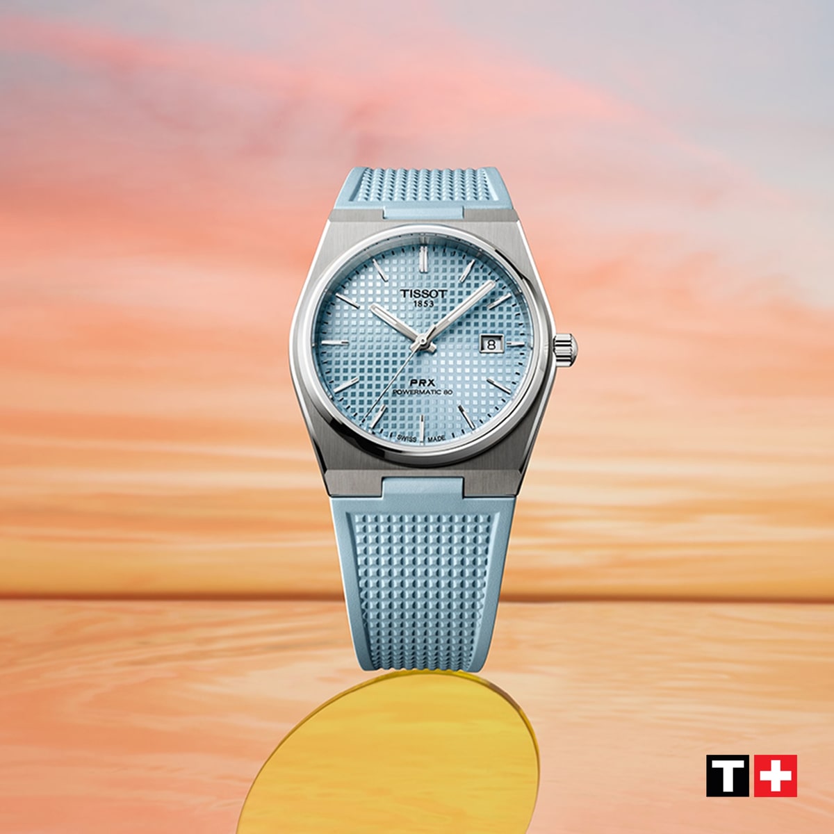 Eine silberne Tissot PRX-Uhr mit einem strukturierten hellblauen Armband und Zifferblatt wird vor einem pastellfarbenen Sonnenuntergangshintergrund dargestellt. Das Tissot-Logo ist in der unteren rechten Ecke zu sehen.