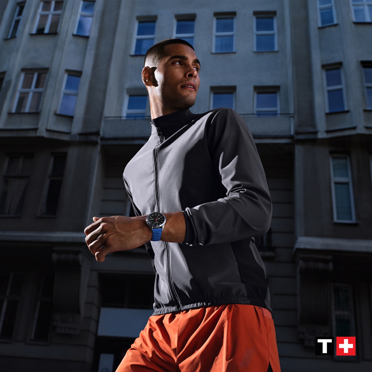 Ein Mann in einer grauen Jacke und orangefarbenen Shorts steht vor einem hohen Gebäude und blickt zur Seite. Er trägt eine blaue Tissot T-Touch Uhr mit dem Tissot-Logo in der rechten unteren Ecke des Fotos.