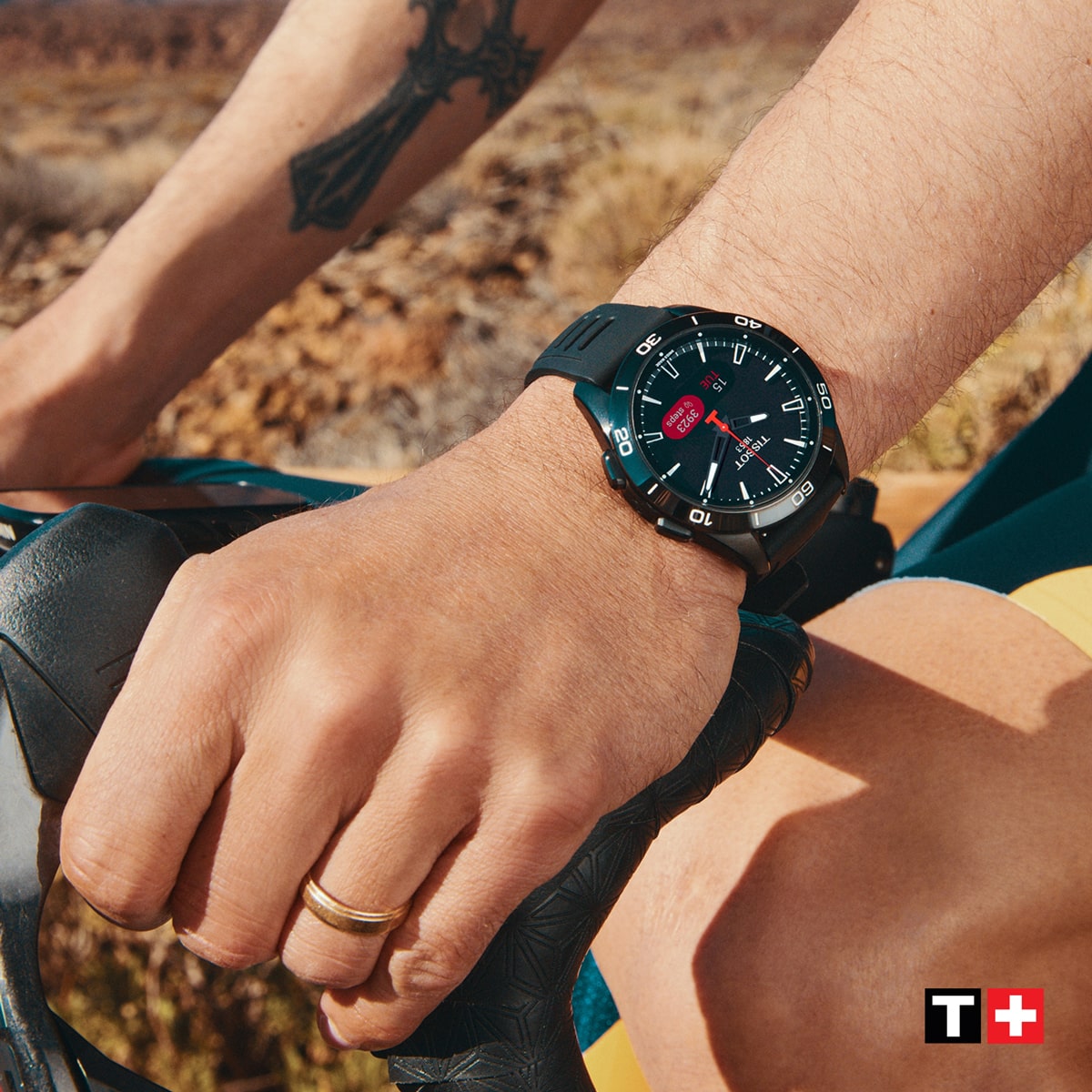 Nahaufnahme der Hand einer Person, die im Freien einen Fahrradlenker umgreift und eine schwarze Tissot T-Touch Armbanduhr trägt. Ein goldener Ring und ein Tattoo betonen den Look, das Tissot-Logo ist in der rechten unteren Ecke zu sehen.