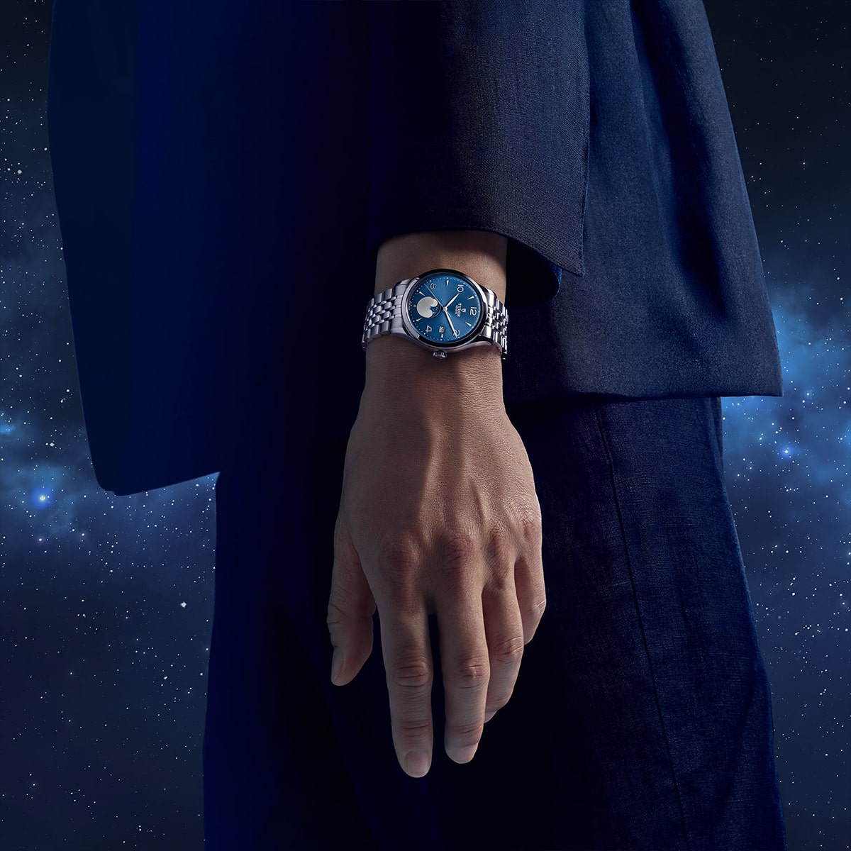 2025 © TUDOR Eine Person in einem dunkelblauen Anzug steht vor dem Hintergrund eines nächtlichen Sternenhimmels und präsentiert eine silberne Armbanduhr von Tudor 1926 mit einem auffallend blauen Zifferblatt an ihrem linken Handgelenk.