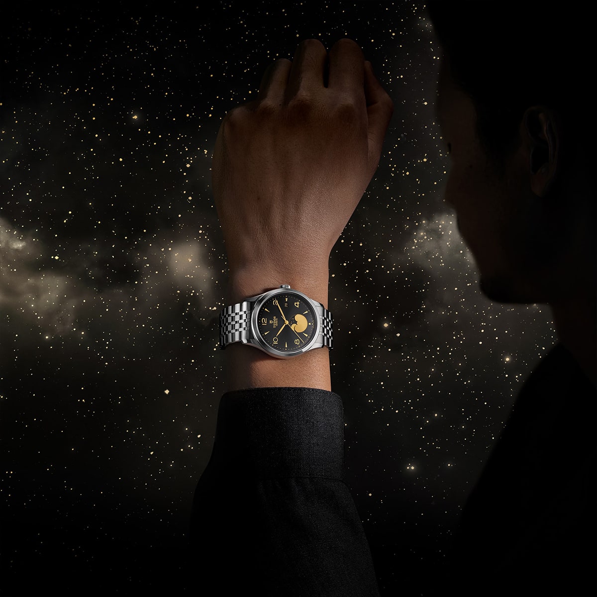 2025 © TUDOR Eine Person, die eine silberne Armbanduhr von Tudor 1926 mit schwarz-goldenem Zifferblatt trägt, streckt ihre Hand vor einem sternenübersäten Nachthimmel aus. Das Zifferblatt zeigt himmlische Motive, die perfekt zum kosmischen Thema passen.