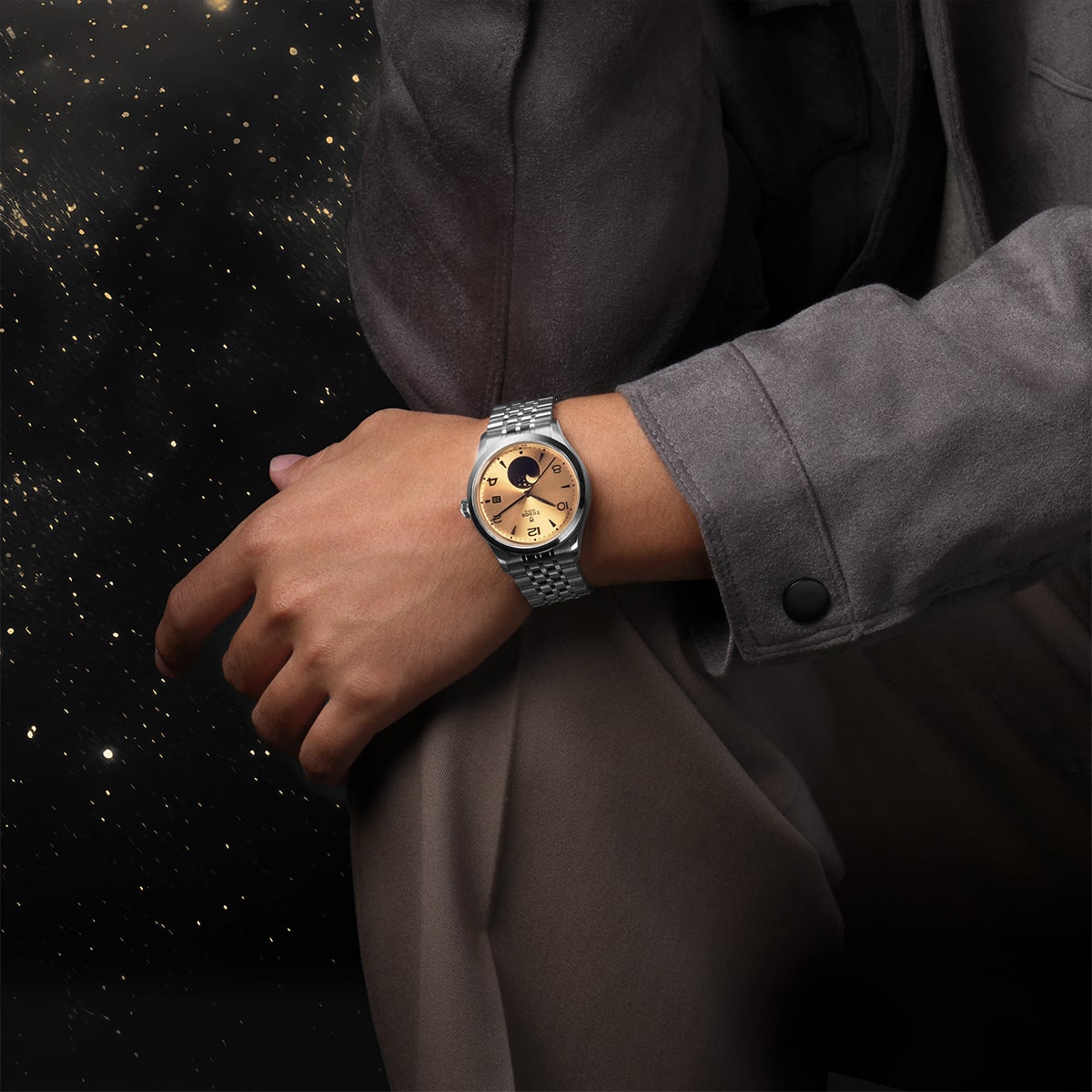 2025 © TUDOR Eine Person, die eine graue Jacke und eine braune Hose trägt, sitzt mit einer Hand auf dem Knie und präsentiert eine goldene und silberne Armbanduhr von Tudor 1926. Im Hintergrund ist ein nächtlicher Sternenhimmel zu sehen.
