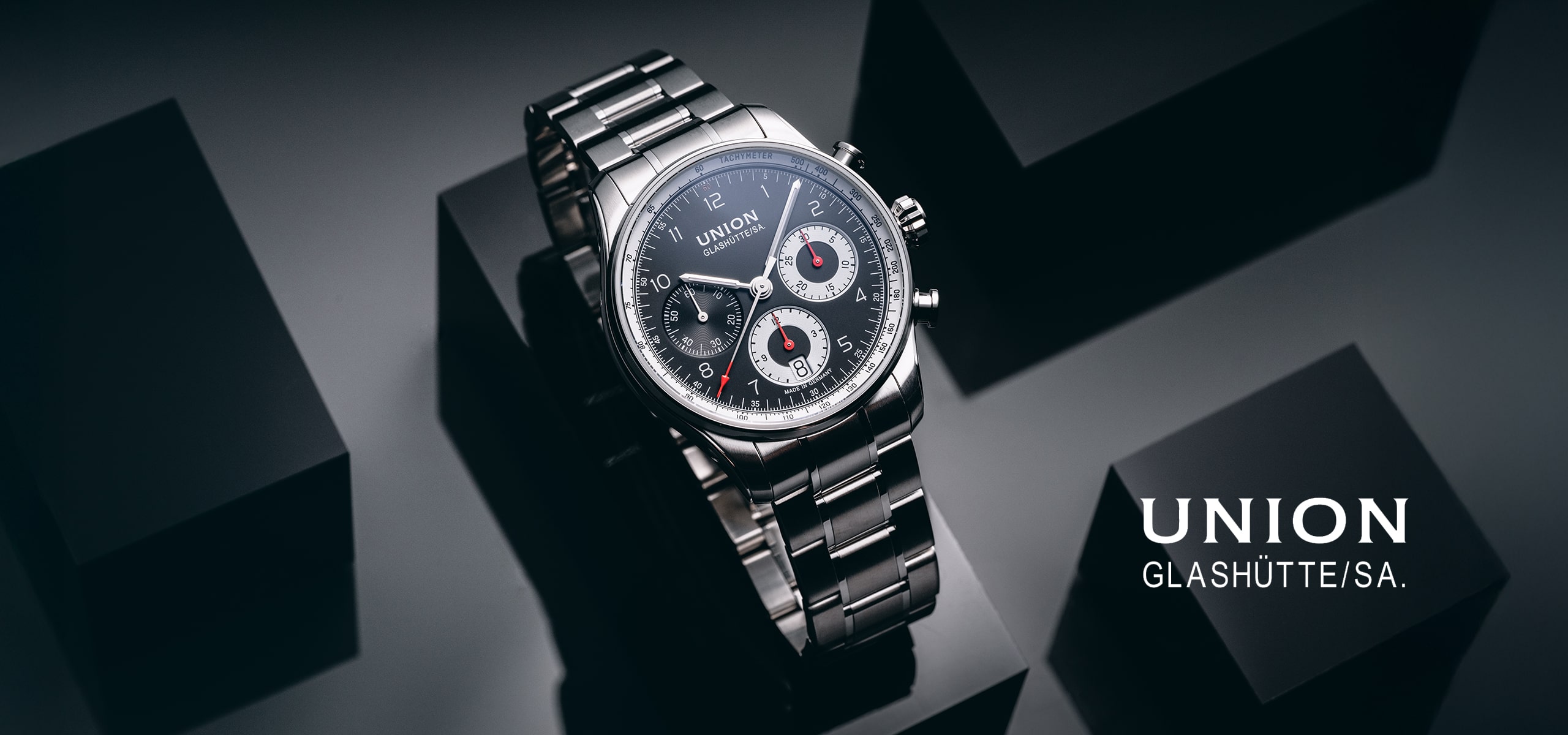 Eine raffinierte Chronographen-Armbanduhr von Union Glashütte wird vor einem eleganten, dunklen geometrischen Hintergrund präsentiert, auf dem rechts in elegantem Weiß "UNION GLASHÜTTE/SA." steht.