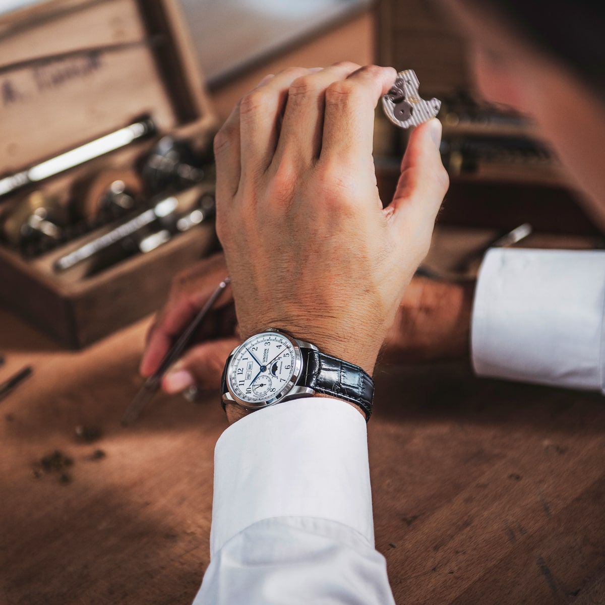 Eine Person in einem weißen Hemd prüft ein Uhrenteil an einer hölzernen Werkbank. Sie trägt eine silber-schwarze Union Glashütte 1893 Armbanduhr. Im Hintergrund ist eine Holzkiste mit Werkzeugen zu sehen.