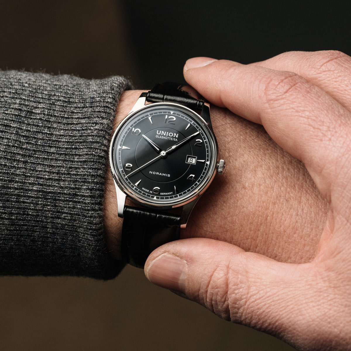 Eine Person, die einen grauen Pullover trägt, stellt eine Union Glashütte Armbanduhr mit schwarzem Lederarmband, rundem schwarzen Zifferblatt und silbernem Gehäuse ein. Der elegante Zeitmesser zeigt die Zeit 10:11 an.