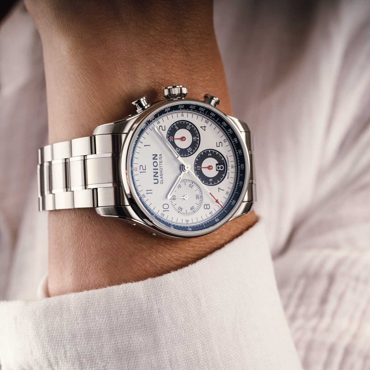 Nahaufnahme einer Person, die eine silberne Chronographen-Armbanduhr Union Glashütte Belisar aus Edelstahl mit weißem Zifferblatt, schwarzen und roten Akzenten und drei Hilfszifferblättern trägt, bekleidet mit einem weißen langärmeligen Hemd.