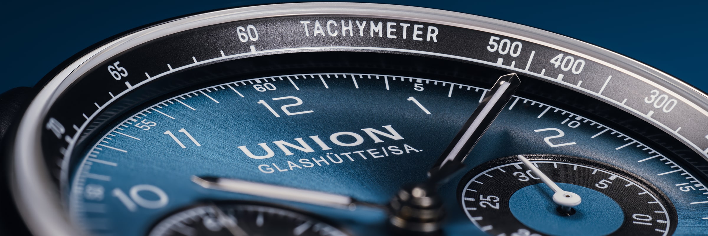 Nahaufnahme einer blauen Union Glashütte Belisar-Armbanduhr, die das Zifferblatt, das Chronographenzifferblatt, die drei Zeiger, die Ziffern und die Tachymeterskala auf der schwarzen Lünette vor blauem Hintergrund hervorhebt.