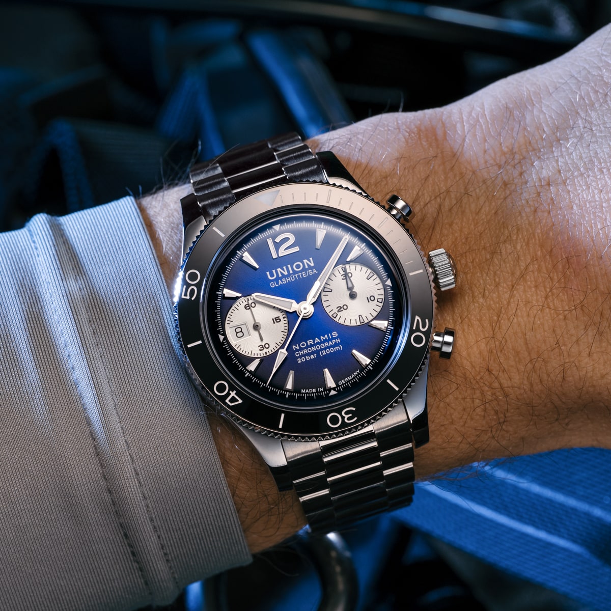 Die Nahaufnahme eines Handgelenks zeigt die Chronographenuhr Union Glashütte Noramis mit blauem Zifferblatt, silbernen Hilfszifferblättern und einem Edelstahlarmband. Die Trägerin kombiniert sie elegant mit einem hellblauen Hemd.