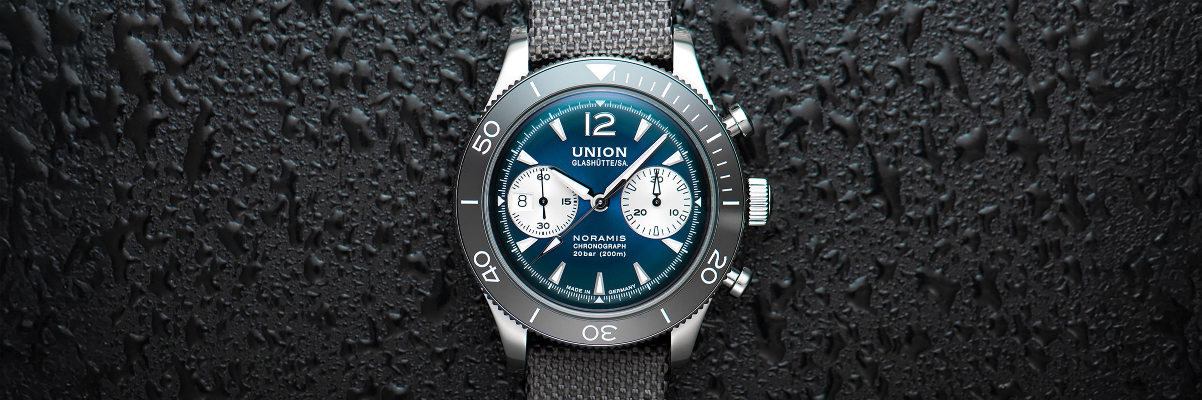 Eine Nahaufnahme einer Union Glashütte Noramis Chronographen-Armbanduhr mit grauem Mesh-Armband, blauem Zifferblatt und weißen Ziffernblättern auf einer strukturierten schwarzen Oberfläche unterstreicht den charakteristischen Stil der Union Glashütte Noramis.