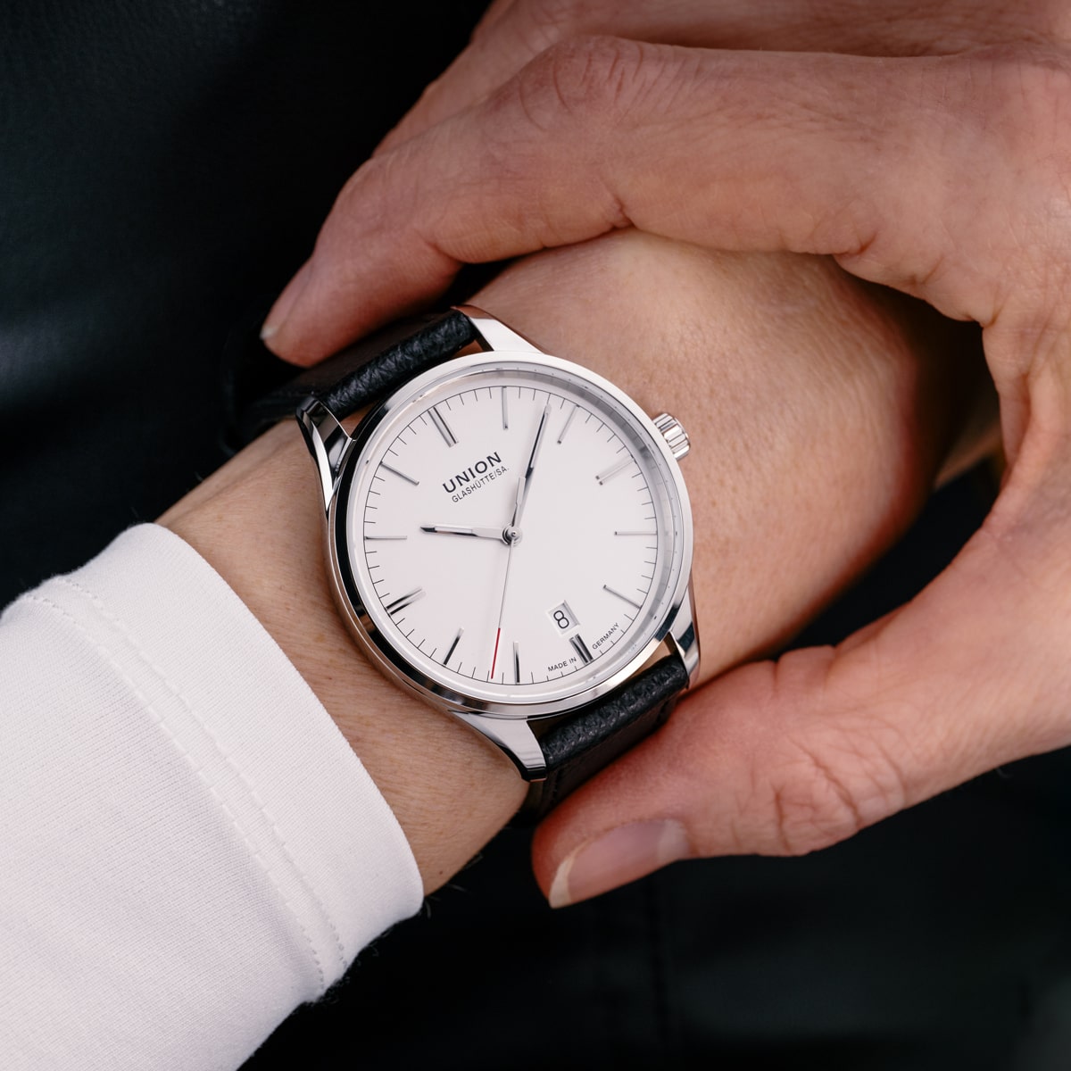 2025 © UNION GLASHÜTTE Eine Person trägt eine klassische silberne Union Glashütte Viro-Armbanduhr mit weißem Zifferblatt, schwarzem Lederarmband, einfachen Stundenmarkierungen und einem Datumsfenster bei 3 Uhr. Eine andere Hand hält sanft das Handgelenk in der Nähe der Uhr.