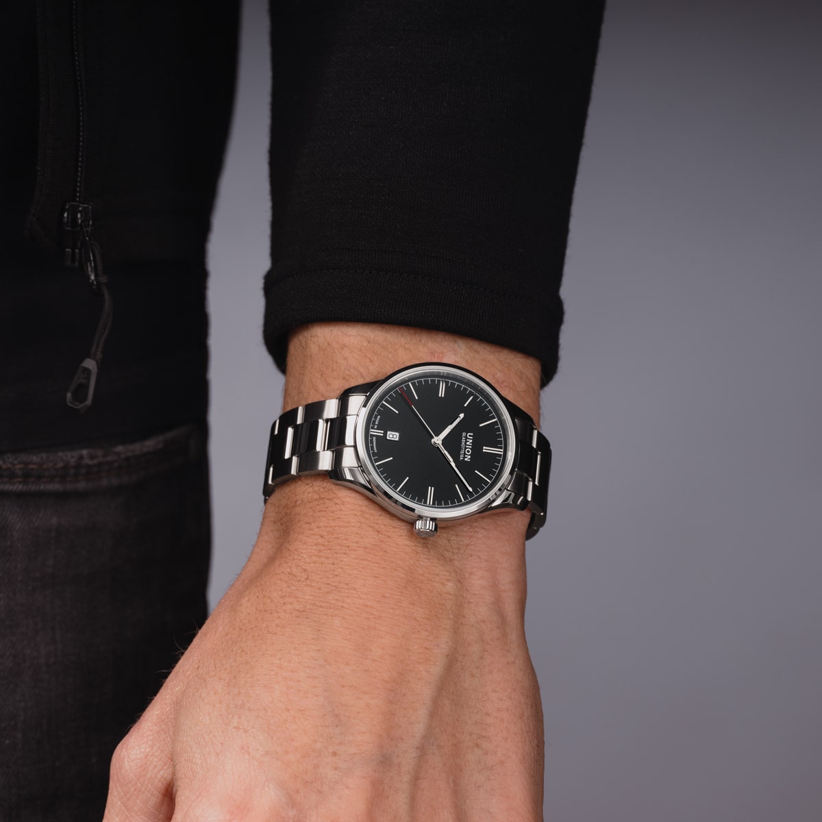 2025 © UNION GLASHÜTTE Eine Person, die ein schwarzes langärmeliges Oberteil und eine analoge Armbanduhr Union Glashütte Viro mit schwarzem Zifferblatt und silbernen Zeigern trägt, steht vor einem schlichten grauen Hintergrund.