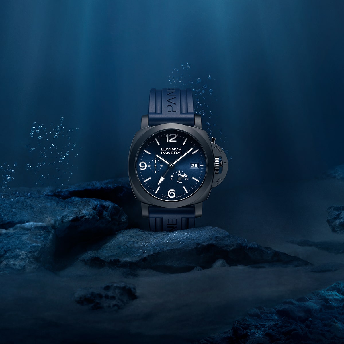 2025 © Panerai Eine dunkelblaue Panerai Luminor Armbanduhr mit Kautschukarmband ruht unter Wasser auf einer felsigen Oberfläche, während Lichtstrahlen durchdringen und Blasen im Hintergrund aufsteigen und die für Panerai typische Eleganz und Beständigkeit zeigen.