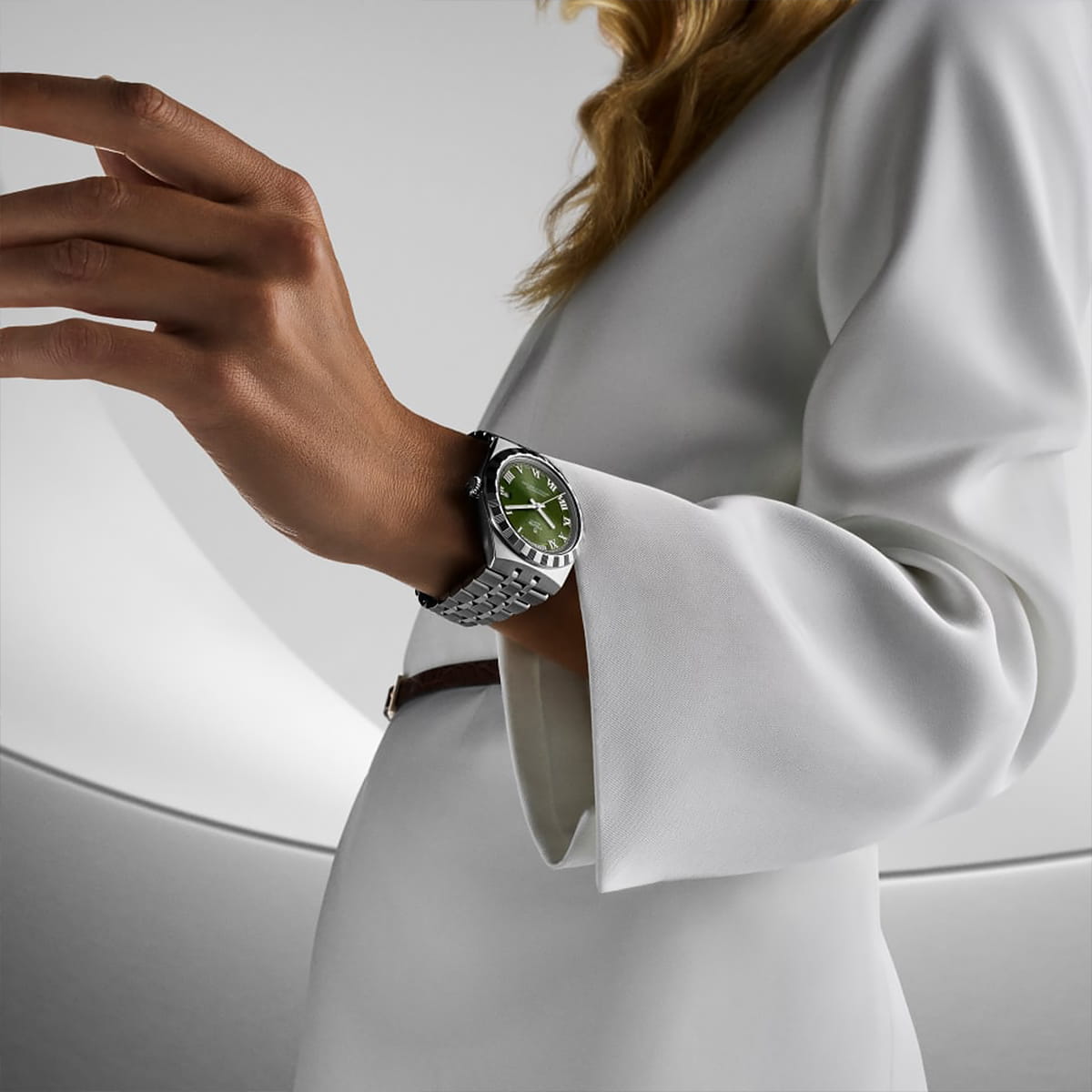 Eine Person in einem weißen, langärmeligen Outfit präsentiert eine silberne TUDOR Royal-Armbanduhr mit grünem Zifferblatt vor einem minimalistischen, hellen Hintergrund.